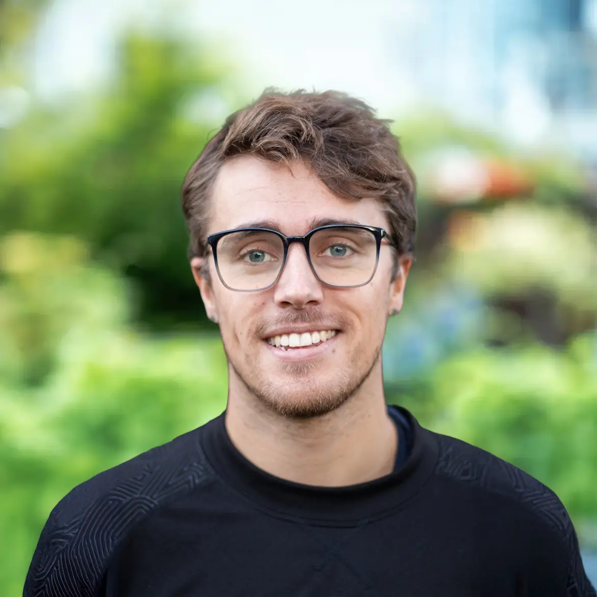 A young man with light brown hair, glasses, and a short beard smiles at the camera while standing outdoors, with greenery blurred in the background, giving a warm glimpse about his cheerful personality.